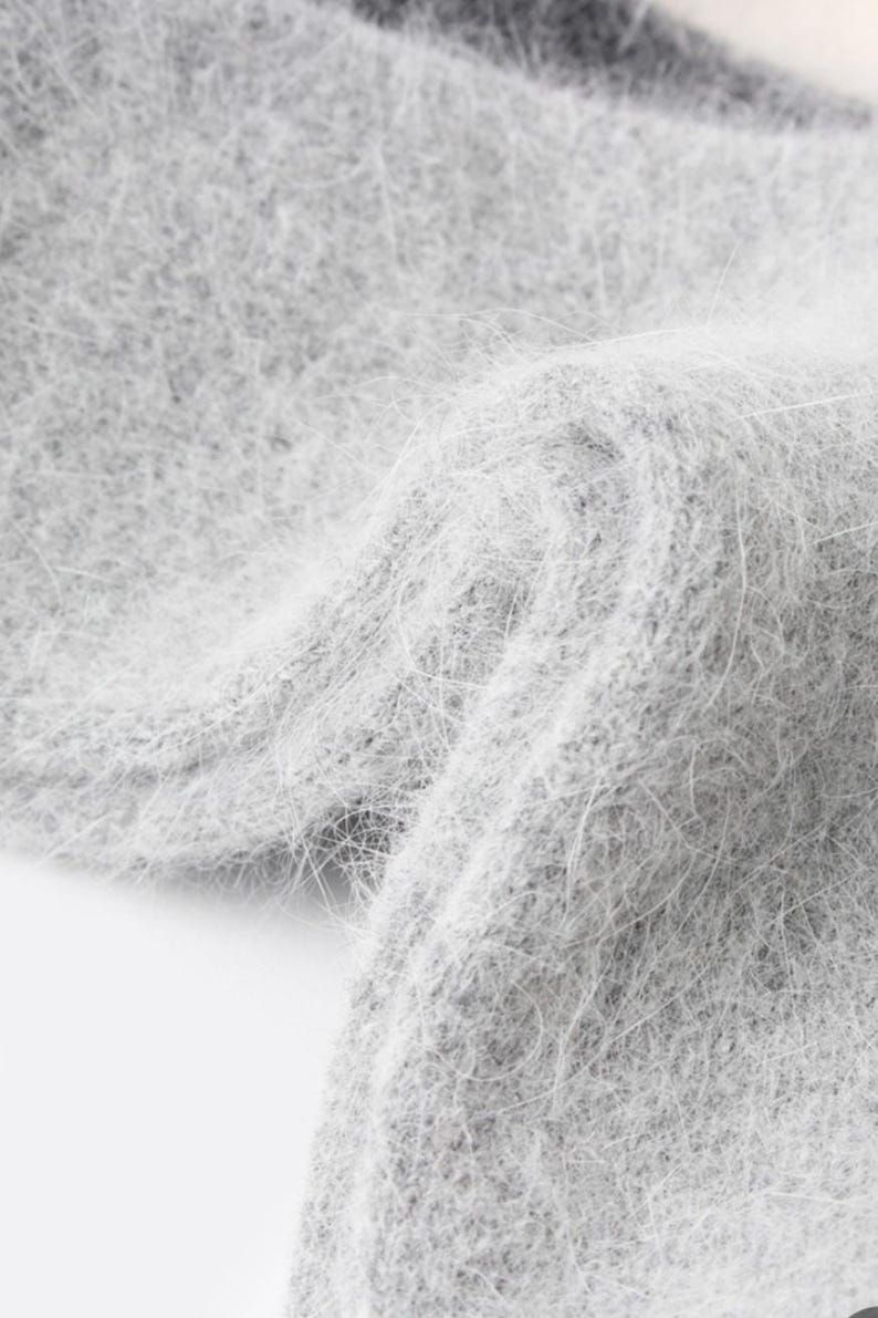 May include: A light gray knit fabric with a soft, fuzzy, and brushed texture. The material features a dense, fibrous surface with long, fine hairs creating a halo effect across the light gray color, showing a folded, layered structure.