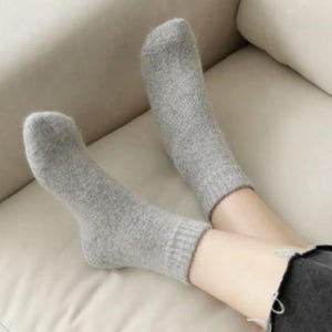 May include: A pair of light gray ankle socks with a soft, fuzzy texture and ribbed cuffs. The socks are worn on the feet, extending just above the ankle, and feature a simple, solid-colored design with a consistent knit pattern throughout.