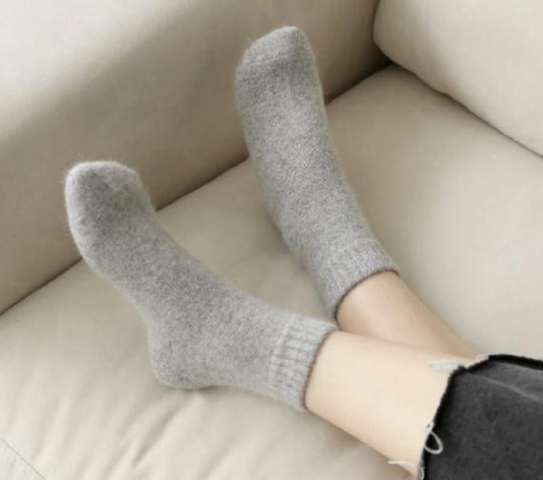 May include: A pair of light gray ankle socks with a soft, fuzzy texture and ribbed cuffs. The socks are worn on the feet, extending just above the ankle, and feature a simple, solid-colored design with a consistent knit pattern throughout.