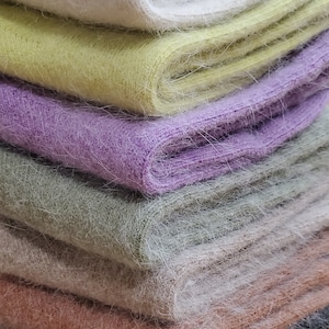 May include: A stack of folded knit scarves featuring a fuzzy, brushed texture. The colors from top to bottom are cream, pale yellow, lavender, sage green, beige, dusty rose, and charcoal gray, all displaying a soft, fibrous appearance.