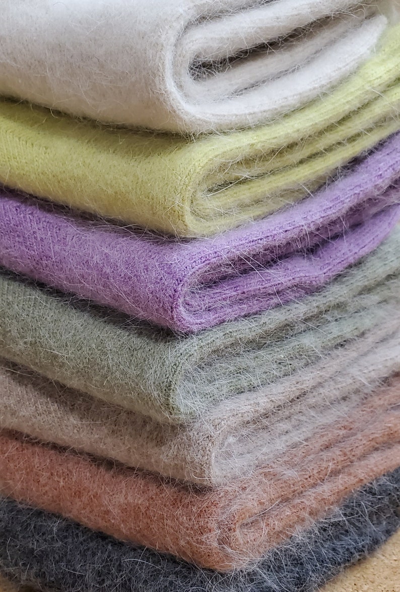 May include: A stack of folded knit scarves featuring a fuzzy, brushed texture. The colors from top to bottom are cream, pale yellow, lavender, sage green, beige, dusty rose, and charcoal gray, all displaying a soft, fibrous appearance.
