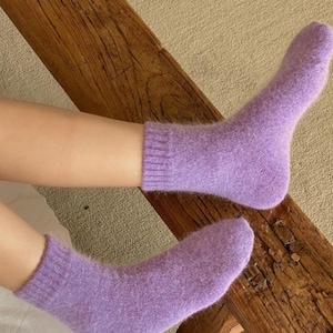 May include: A pair of ankle-length socks in a soft, fuzzy lavender knit material. The socks feature a ribbed cuff at the top and a solid, uniform light purple color throughout, providing a textured appearance with a visible soft pile.