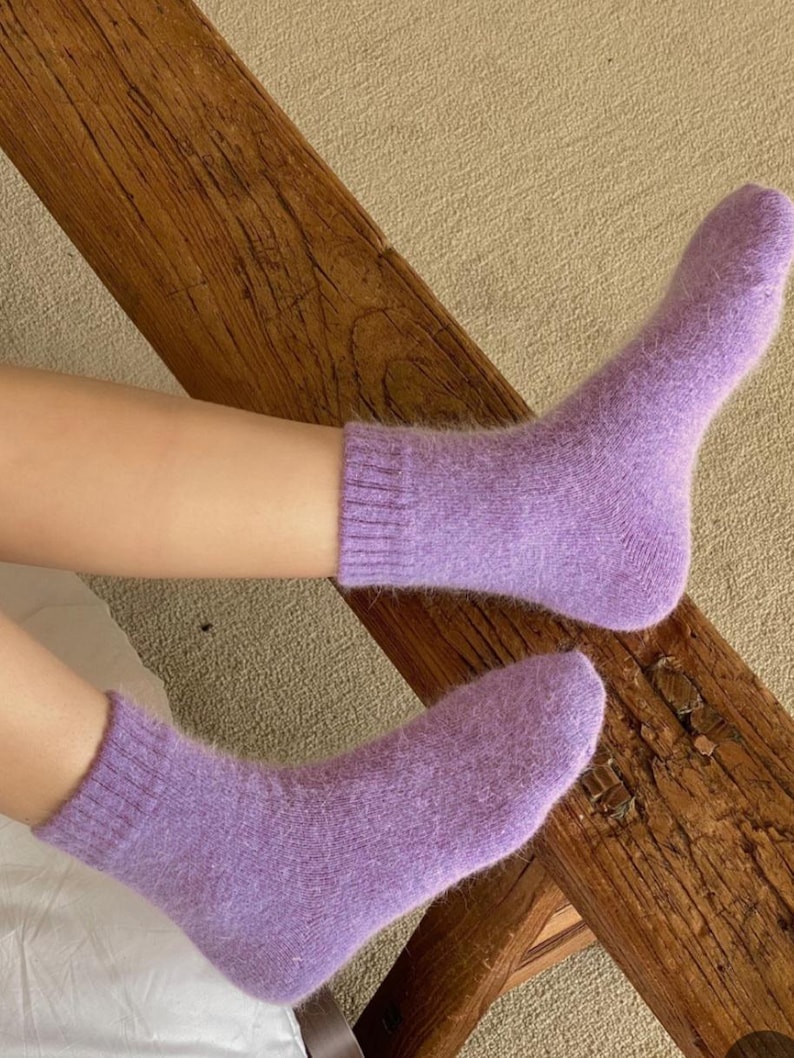 May include: A pair of ankle-length socks in a soft, fuzzy lavender knit material. The socks feature a ribbed cuff at the top and a solid, uniform light purple color throughout, providing a textured appearance with a visible soft pile.