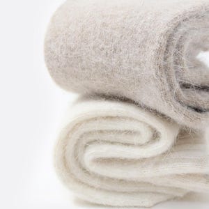 May include: A pair of folded socks featuring a fuzzy, brushed texture. The top sock is a light beige or oatmeal color, while the bottom sock is a solid off-white. Both items exhibit a soft, fibrous appearance with visible knit loops.