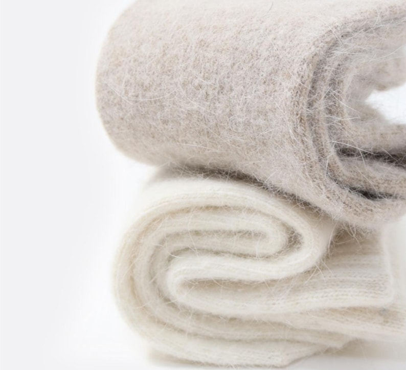 May include: A pair of folded socks featuring a fuzzy, brushed texture. The top sock is a light beige or oatmeal color, while the bottom sock is a solid off-white. Both items exhibit a soft, fibrous appearance with visible knit loops.