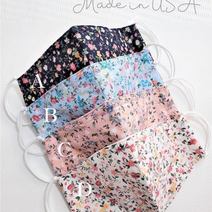 May include: Four floral print face masks with nose wire and filter pocket. The masks are layered in a stack, with the top mask being black with white and pink flowers, the second mask is light blue with white and pink flowers, the third mask is pink with white and pink flowers, and the bottom mask is white with pink and yellow flowers. The text "Ready to shop Made in USA" is at the top of the image. The text "Nose Wire + Filter Pocket" is at the bottom of the image.