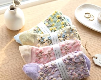 Op de afbeelding: Vier paar bloemen geborduurde sokken in pastelkleuren: groen, beige, roze en paars. Elk paar is verpakt met een witte band. De sokken zijn gerangschikt op een houten oppervlak in de buurt van een raam, met een kleine vaas en decoratieve items op de achtergrond.