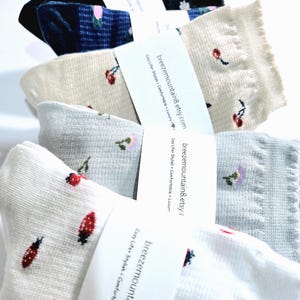 Puede incluir: Un montón de cinco pares de calcetines con diferentes patrones florales y de cerezas. Los calcetines son blancos, beige, azul claro y azul marino. Los calcetines están etiquetados con el texto "breezemountain8.etsy.com" y "Cozy Life Stylish Comfortable Luxury".