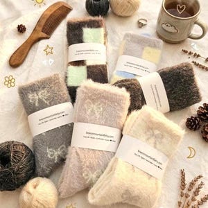 May include: A collection of cozy socks in various colors and patterns, including checkered and bow designs. The socks are bundled with white paper bands. The image also includes yarn balls, a wooden comb, and a mug of tea.