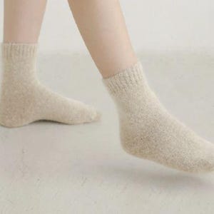 May include: A pair of ankle-length socks in a light beige, heathered knit texture. The socks feature a ribbed cuff at the top and a soft, fuzzy appearance, covering the feet and extending slightly above the ankle bone in a neutral tone.
