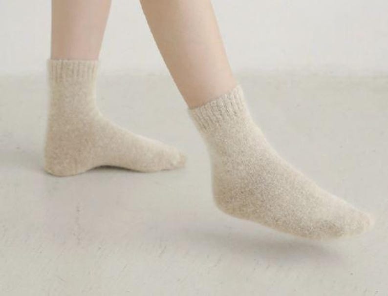 May include: A pair of ankle-length socks in a light beige, heathered knit texture. The socks feature a ribbed cuff at the top and a soft, fuzzy appearance, covering the feet and extending slightly above the ankle bone in a neutral tone.