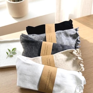 May include: A stack of five pairs of socks in various colors: black, gray, charcoal, beige, and white. Each pair is folded and wrapped with a brown paper band. The socks have a ruffled edge.