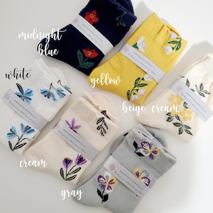 May include: Assortment of floral patterned socks in various colors, including white, cream, gray, beige, yellow, and midnight blue. Each sock features a different floral design and is folded in half. The socks are arranged in a circle on a white surface. The text "www.breezemountainetsy.com" and "thank you so much for your purchase" is printed on a tag attached to each sock.