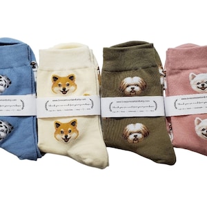 May include: Four pairs of dog-themed socks in various colors: light blue, cream, olive green, and pink. Each sock features an embroidered dog face. The socks are wrapped with a white band that includes text.
