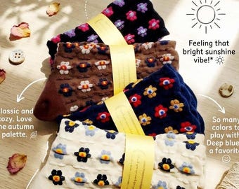 Op de afbeelding: Vier paar sokken met patronen in zwart, bruin, marineblauw en crème, elk met een bloemenmotief. De sokken zijn samengebonden met een geel lint. De afbeelding bevat tekst die luidt "Luxe texture & cool pattern!" en "Feeling that bright sunshine vibe!"