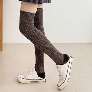 Op de afbeelding: Bruine gebreide dijbeenwarmers. De beenwarmers worden gedragen met witte sneakers met zwarte accenten. De beenwarmers zijn geribbeld en hebben een gespikkelde textuur. De achtergrond is een effen witte muur en een lichtgekleurde vloer.