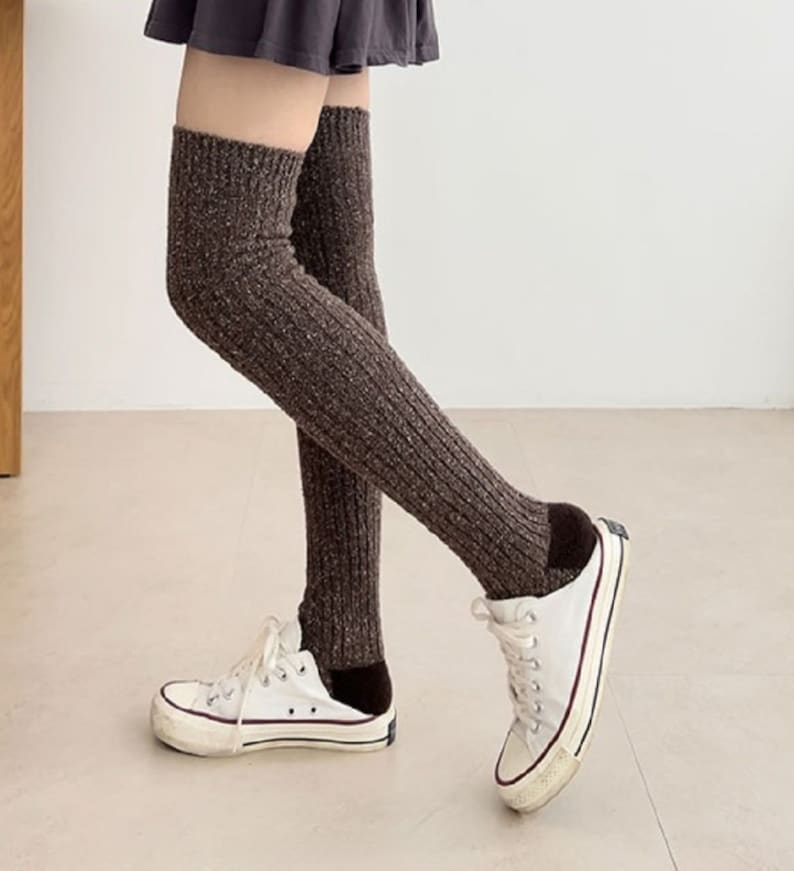 Op de afbeelding: Bruine gebreide dijbeenwarmers. De beenwarmers worden gedragen met witte sneakers met zwarte accenten. De beenwarmers zijn geribbeld en hebben een gespikkelde textuur. De achtergrond is een effen witte muur en een lichtgekleurde vloer.