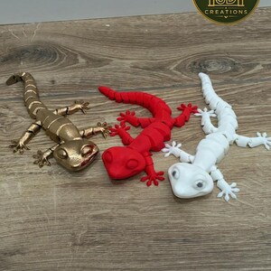 Op de afbeelding: Drie gearticuleerde hagedisbeeldjes in metallic goud, matrood en matwit. Elke hagedis heeft een gesegmenteerd lichaam, vier poten met gespreide tenen en een gedetailleerde kop. Een rond logo rechtsboven toont MF 3D CREATIONS.