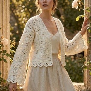 Puede incluir: Un cárdigan de ganchillo color blanco roto con un diseño de punto de cuadrados, mangas largas acampanadas y un dobladillo festoneado. La prenda se lleva sobre una sencilla blusa sin mangas de color claro y una falda a juego, con texturas.