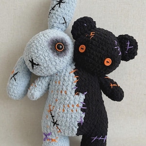 Può includere: Un peluche dal design diviso, con un lato a forma di coniglio azzurro e l'altro di orso nero. Il coniglio presenta una macchia scura intorno all'occhio e cuciture nere, mentre l'orso ha occhi a bottone arancioni e punti viola e arancioni.