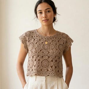 May include: A light brown crochet top featuring a repeating floral motif pattern. The garment has short sleeves, a round neckline, and a scalloped hemline. The intricate needlework creates a textured, openwork design throughout the entire piece.