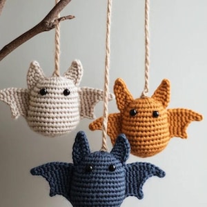 Puede incluir: Tres adornos de murciélago tejidos a ganchillo que cuelgan de una rama mediante cordeles. Cada murciélago tiene un cuerpo redondeado, orejas puntiagudas y ojos de cuentas negras. Son de color beige claro, naranja dorado y azul oscuro, con texturas.