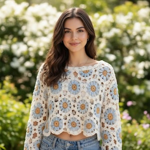 May include: A long-sleeved crochet sweater featuring a repeating pattern of daisy-like floral motifs. The flowers have light blue petals and mustard yellow centers, connected by cream-colored yarn. The garment has a scalloped hem and neckline.