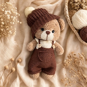 Könnte beinhalten: Ein gehäkelter Teddybär, der eine dunkelbraune Mütze mit einem cremefarbenen Bommel und eine passende dunkelbraune Latzhose mit zwei kleinen Holzknöpfen trägt. Der Bär hat hellbraunes Fell, eine cremefarbene Schnauze und dunkelbraune Augen.