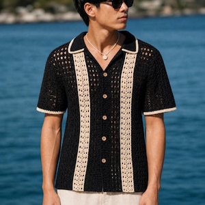May include: A short-sleeved, black crochet button-down shirt featuring a collared neckline and two vertical beige crochet panels running down the front. The shirt has a textured, open-knit pattern throughout and is finished with beige trim on the sleeves.