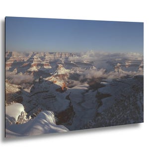 May include: A rectangular wall art print featuring a panoramic view of the Grand Canyon covered in a layer of white snow. The landscape displays deep red rock formations partially obscured by low-hanging white clouds under a clear blue sky.
