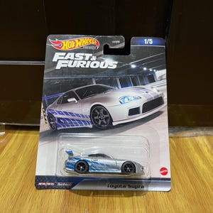 Może przedstawiać: Model samochodu Hot Wheels Premium Fast & Furious Toyota Supra w kolorze srebrnym z niebieską grafiką na bokach, zamknięty w oryginalnym opakowaniu typu blister. Karta zawiera logo Fast & Furious, napis 1/5 oraz grafikę samochodu.