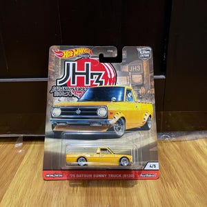 Peut inclure: Un camion miniature Hot Wheels Premium 1975 Datsun Sunny Truck (B120) jaune, dans son emballage d'origine. La carte présente le texte 'JH3', 'JAPAN HISTORIC' et '4/5', avec une illustration de camion jaune sur un fond de soleil rouge.