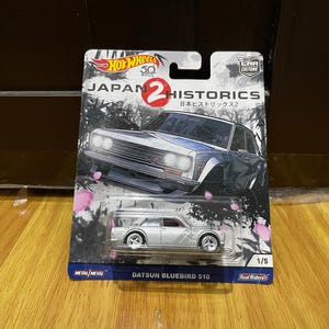 Peut inclure: Une voiture miniature Datsun Bluebird 510 argentée dans son emballage d'origine. La carte présente un graphique de la voiture avec des pétales de cerisier et le texte Hot Wheels 50, Japan Historics 2 et Datsun Bluebird 510.