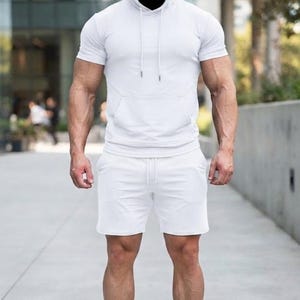 May include: A solid white short-sleeved hooded sweatshirt featuring a front kangaroo pocket and matching white drawstring cords, paired with solid white athletic shorts that have an elastic waistband and a drawstring closure.