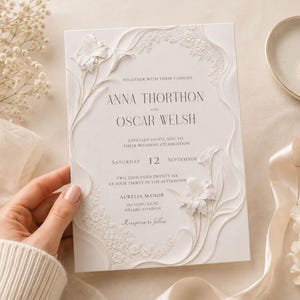 May include: A white wedding invitation card featuring embossed floral details and elegant black serif typography. The text reads: Together with their families, Anna Thorthon and Oscar Welsh, joyfully invite you to their wedding celebration, Saturday 12 September, two thousand twenty six, at four thirty in the afternoon, Aurelia Manor, 165 Long Lane, London, Reception to follow.