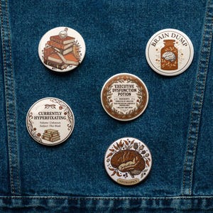 Peut inclure: Cinq badges ronds avec des illustrations et du texte. Les motifs incluent une pile de livres, un cerveau dans un bocal marqué BRAIN DUMP, une étiquette EXECUTIVE DYSFUNCTION POTION, une étiquette CURRENTLY HYPERFIXATING avec des livres et un cerveau marqué NEUROSPICY.