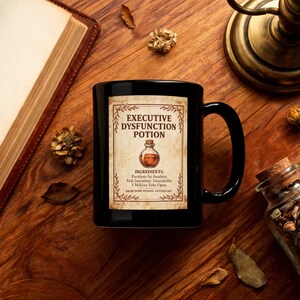 May include: A black ceramic mug featuring a rectangular label with the text Executive Dysfunction Potion, an illustration of a small potion bottle, and a list of ingredients including Paralysis by Analysis and 1 Million Tabs Open.