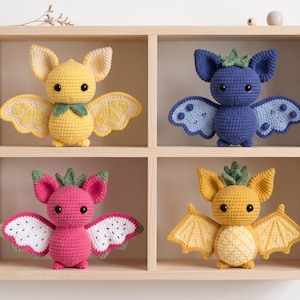 May include: Four crocheted bat plushies in a wooden shelf. Top left is yellow with lemon-slice wings. Top right is blue with berry-like wings. Bottom left is pink with dragon fruit wings. Bottom right is golden yellow with pineapple-textured wings.