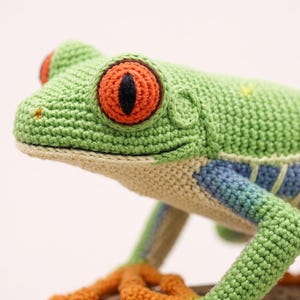May include: A crocheted red-eyed tree frog featuring a bright green back, large orange eyes with black pupils, a cream-coloured underside, and blue and yellow striped flanks. The frog has orange feet and is crafted with visible yarn stitches.