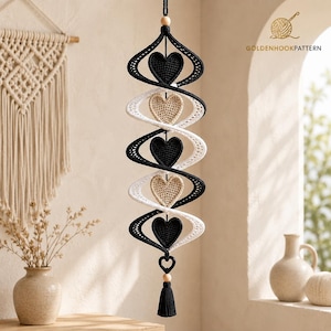 May include: A vertical hanging decoration featuring a spiral structure made of black and white crocheted yarn. The spiral contains five alternating heart-shaped motifs in black and beige. A wooden bead and a black tassel hang from the bottom.