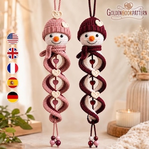May include: Two crocheted snowman ornaments with spiral bodies, one in dusty pink and one in deep burgundy. Each features a matching beanie with a small heart, a carrot nose, and a central string adorned with small hearts and beads. Text reads GOLDENHOOKPATTERN.