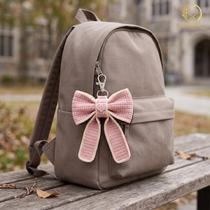 Könnte beinhalten: Ein taupefarbener Rucksack mit einem gehäkelten Schleifen-Schlüsselanhänger am vorderen Reißverschluss. Die Schleife ist altrosa mit einem cremefarbenen Rand und einem kleinen herzförmigen Detail in der Mitte. Der Rucksack hat ein Hauptfach.