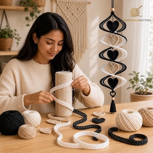 May include: A hanging crochet spiral mobile featuring alternating black and cream-colored hearts. The mobile is suspended by a black cord with a wooden bead and a black tassel at the bottom. Several balls of yarn and loose crochet hearts are present.