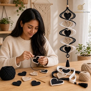 May include: A vertical hanging mobile featuring alternating black and white crocheted hearts connected in a spiral structure, with a black tassel at the bottom. Several loose black and beige crocheted hearts, yarn balls, and a needle are on the table.