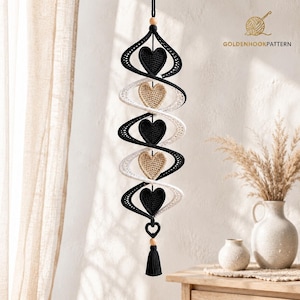 May include: A hanging spiral decoration featuring alternating black and white crocheted ribbons. Five heart-shaped elements are centered within the spiral, alternating between black and beige. The piece includes wooden beads and a black tassel.