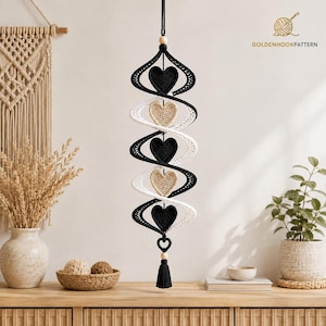 May include: A hanging crochet spiral mobile featuring alternating black and beige heart-shaped ornaments. The structure is suspended by a black cord with wooden beads at the top and bottom, finished with a black tassel. The text GOLDENHOOKPATTERN appears in the upper right corner.