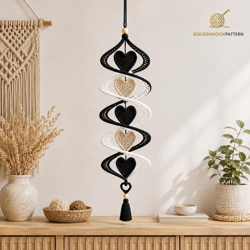 May include: A hanging crochet spiral mobile featuring alternating black and beige heart-shaped ornaments. The structure is suspended by a black cord with wooden beads at the top and bottom, finished with a black tassel. The text GOLDENHOOKPATTERN appears in the upper right corner.