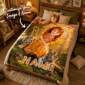 Puede incluir: Una manta de forro polar suave con la imagen impresa de un niño pelirrojo con un vestido amarillo, sentado en un entorno forestal con dos tigres. La manta incluye el texto "NAME" en letras grandes de color naranja y un área personalizada.