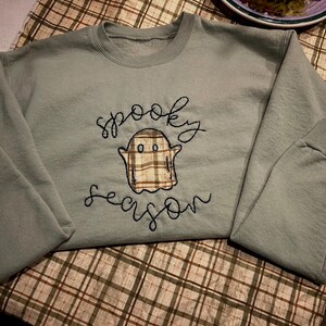 May include: A sage green crewneck sweatshirt featuring an embroidered ghost character with a brown and tan plaid pattern. The words spooky season are stitched in black cursive lettering above and below the ghost graphic on the front chest.