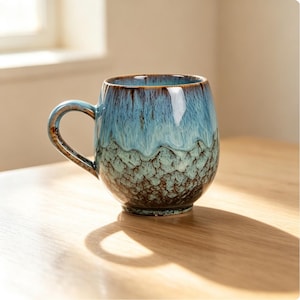 May include: A ceramic mug with a rounded body and a curved handle. The exterior features a reactive glaze with a smooth, glossy light blue top section that transitions into a textured, mottled green and dark brown base with a crackled finish.
